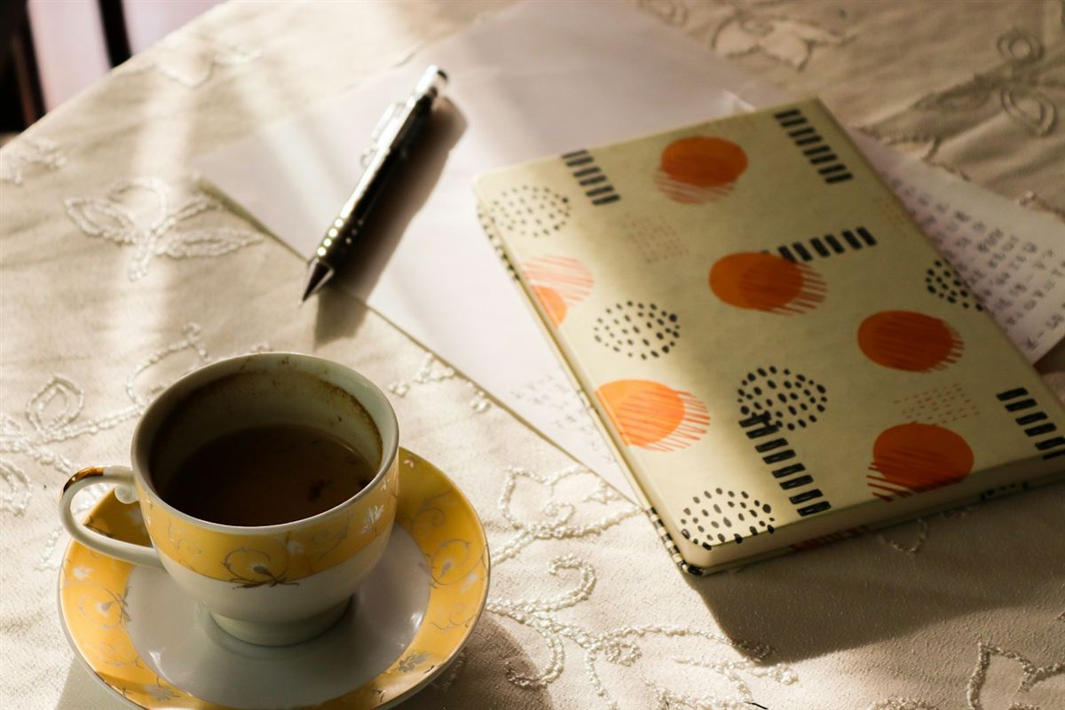 An orange notebook, glasses and a cup of coffee on a sunlit desk